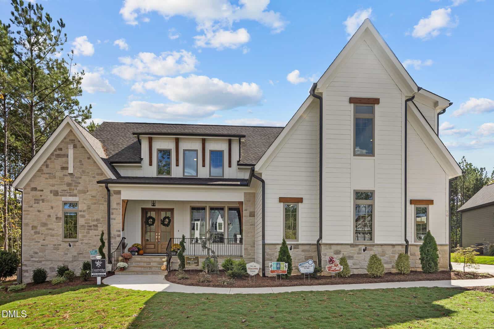 A two-story house with light brick and white siding, black trim windows, a covered front porch with columns, decorative signs on the lawn, and landscaping under a blue sky with scattered clouds.