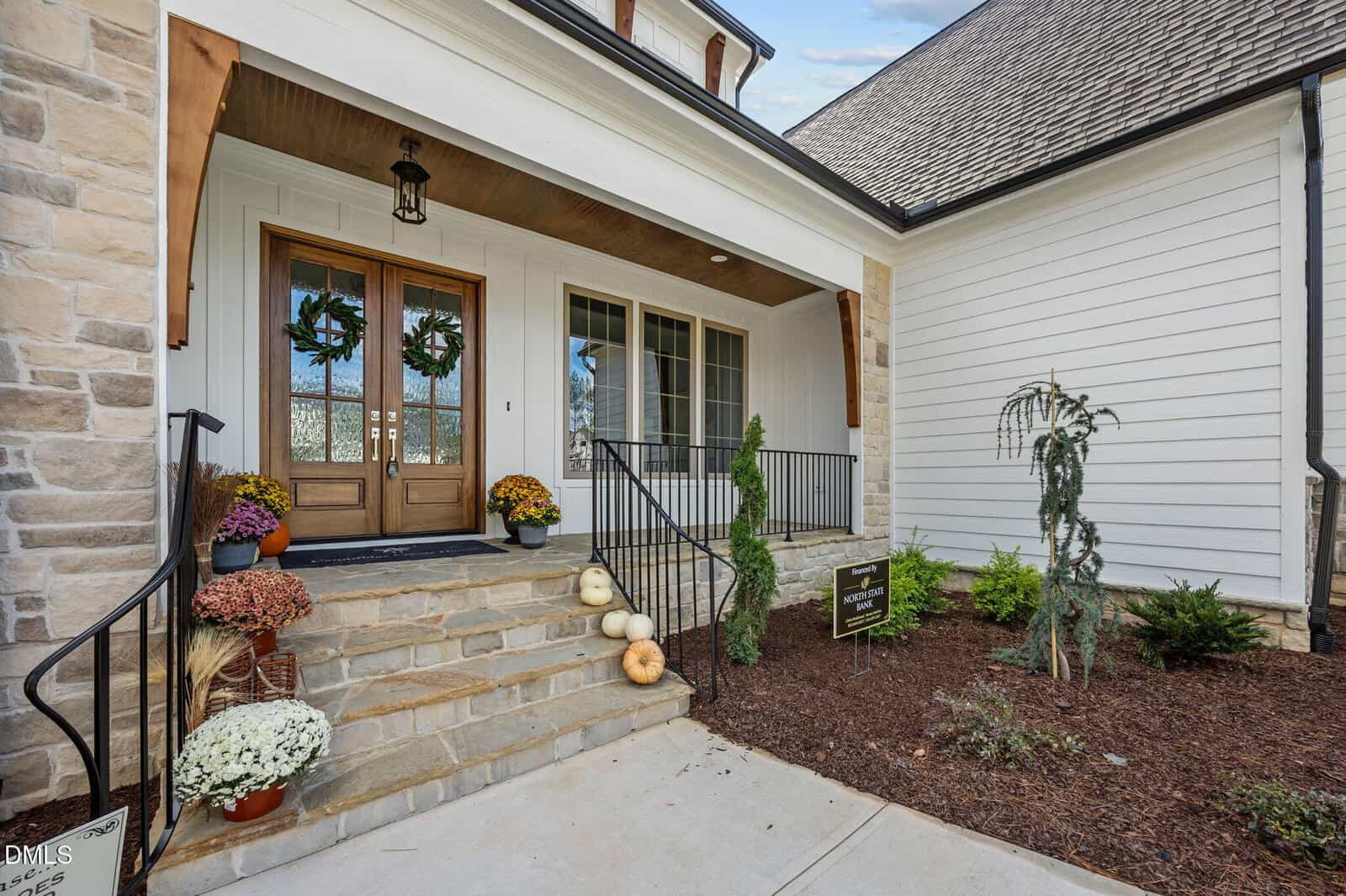 A stone and white-paneled house entrance with double wooden doors, autumn wreaths, potted mums, and stacked pumpkins on the steps. Black railings and minimal landscaping are visible by the porch.