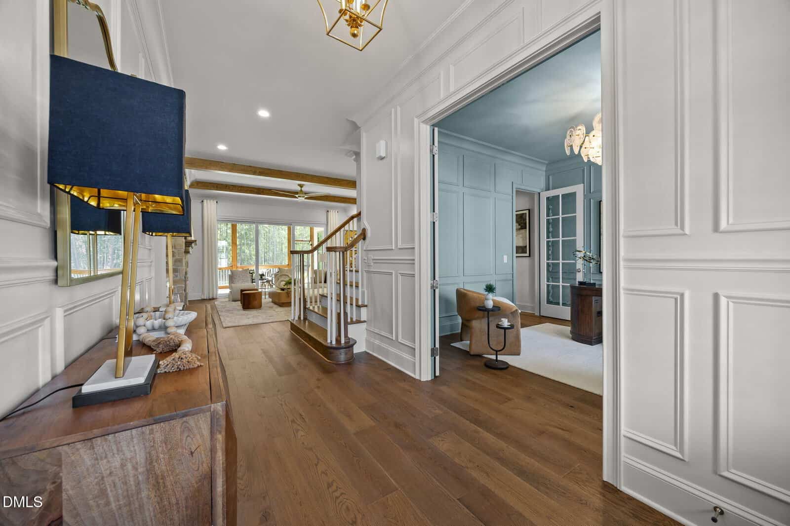 A hallway with light wood floors, white paneled walls, a gold-accented lamp on a console, an open doorway to a sitting room, a staircase, and a view of a living area with large windows in the background.