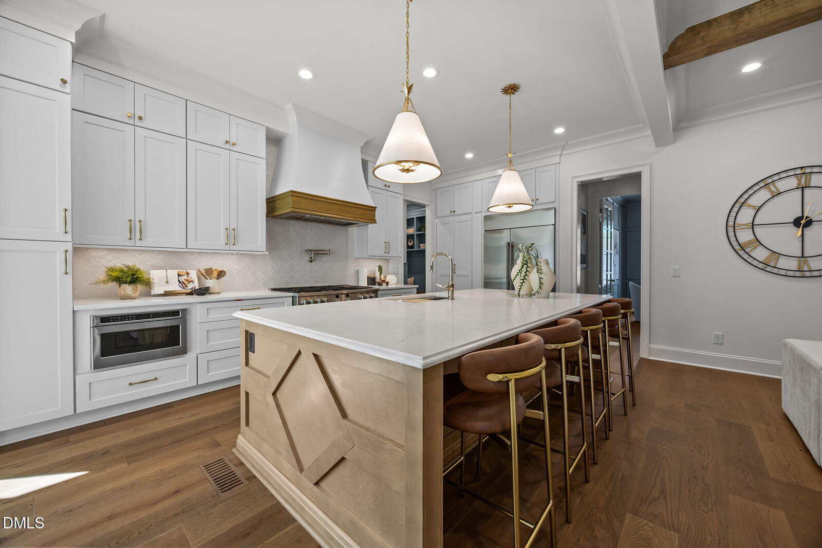 Modern kitchen with white cabinets, a large marble island with gold fixtures, brown leather bar stools, pendant lights, wood flooring, and a wall clock. A microwave and decorative items are on the far counter.