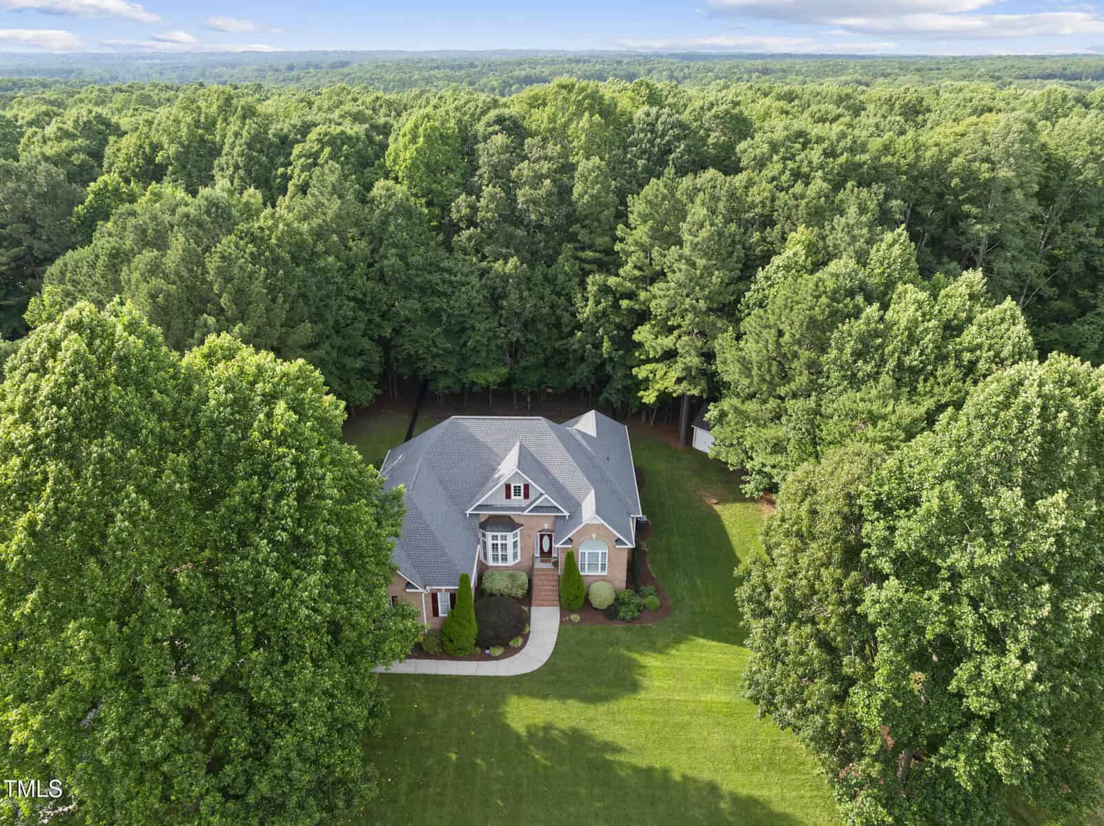 Aerial view of a brick house with a gray roof, surrounded by lush green trees and a large yard, bordered by dense forest under a partly cloudy sky.