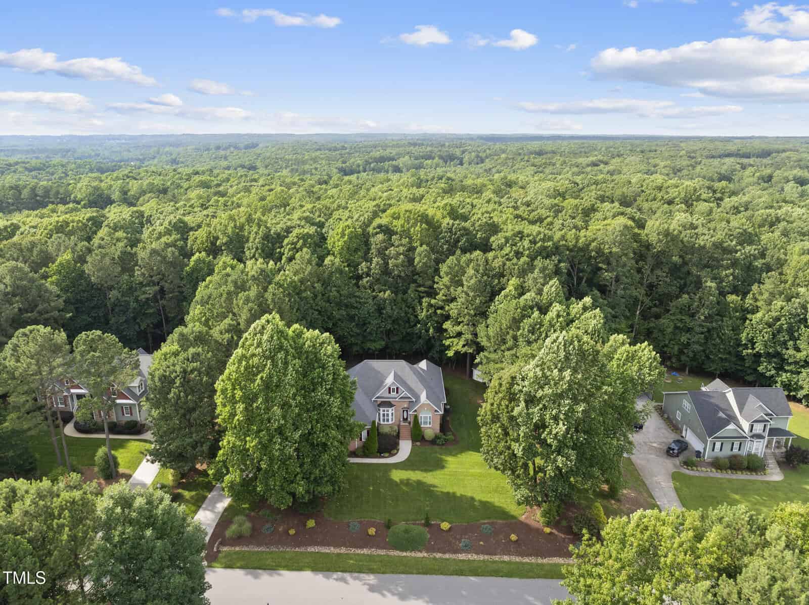 Aerial view of three single-family homes with large front lawns and driveways, surrounded by dense green trees and forest under a blue sky with scattered clouds.