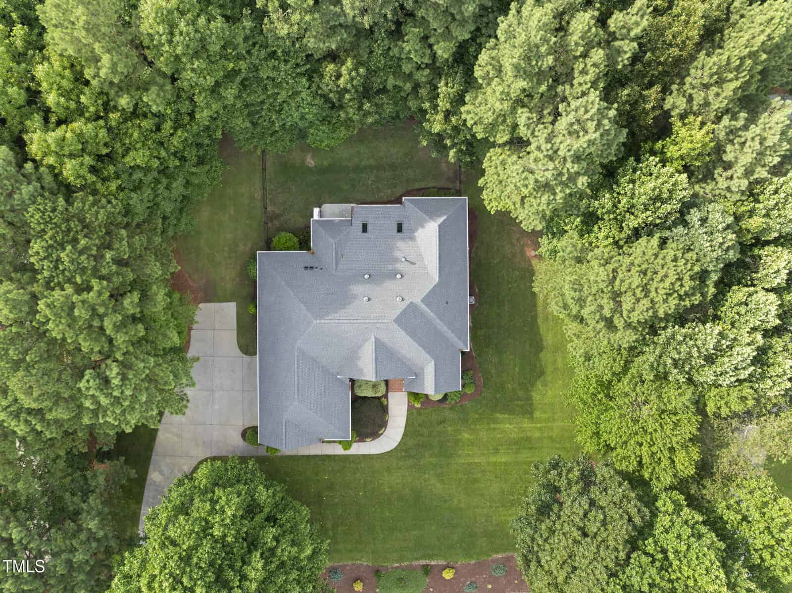 Aerial view of a two-story house with a grey roof, surrounded by trees and green lawn. A driveway leads to a two-car garage on the left side of the house. The property is bordered by dense forest.