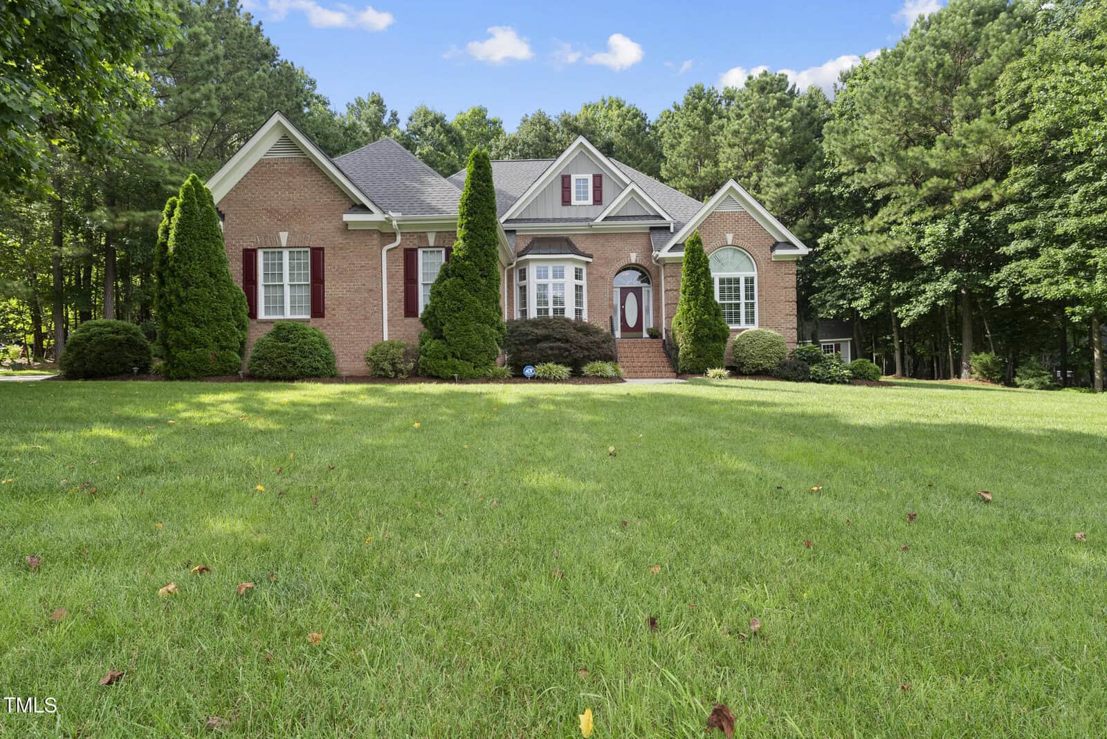A red-brick house with white trim and red shutters is surrounded by tall trees and a large, well-maintained grassy lawn under a blue sky with scattered clouds.