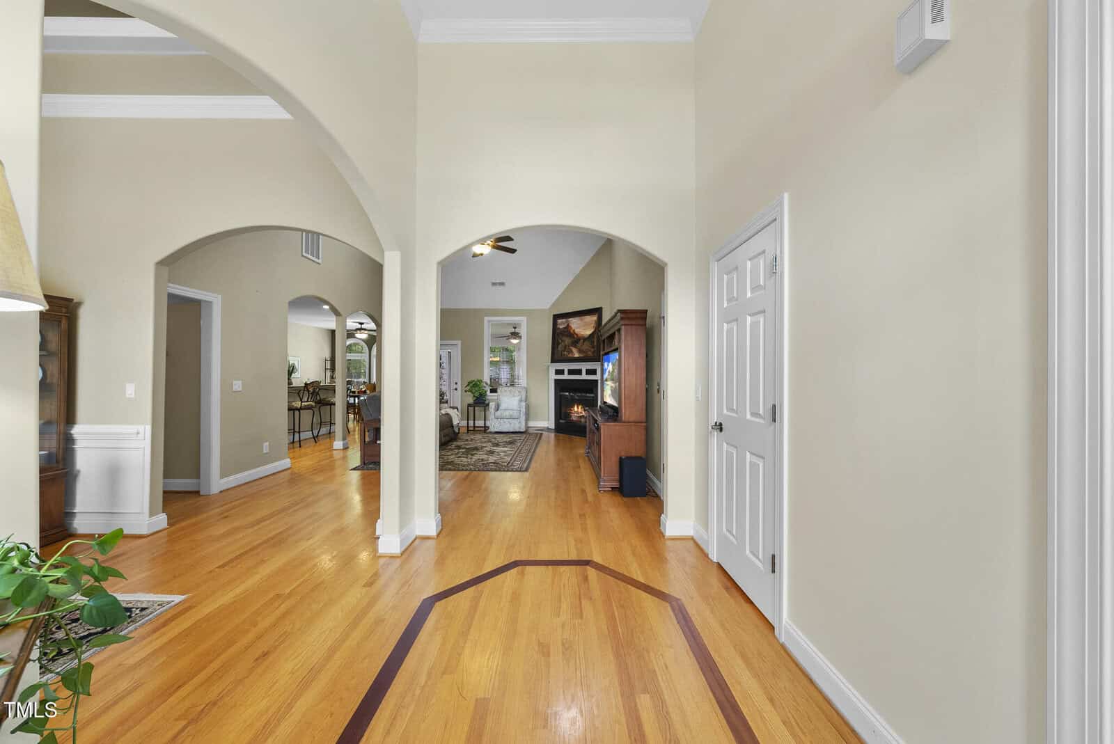 A hallway with wood floors and cream-colored walls leads to a living area with a fireplace. Multiple arched doorways and a white door are visible, along with a plant on the left. Ceiling fan and furniture in the background.