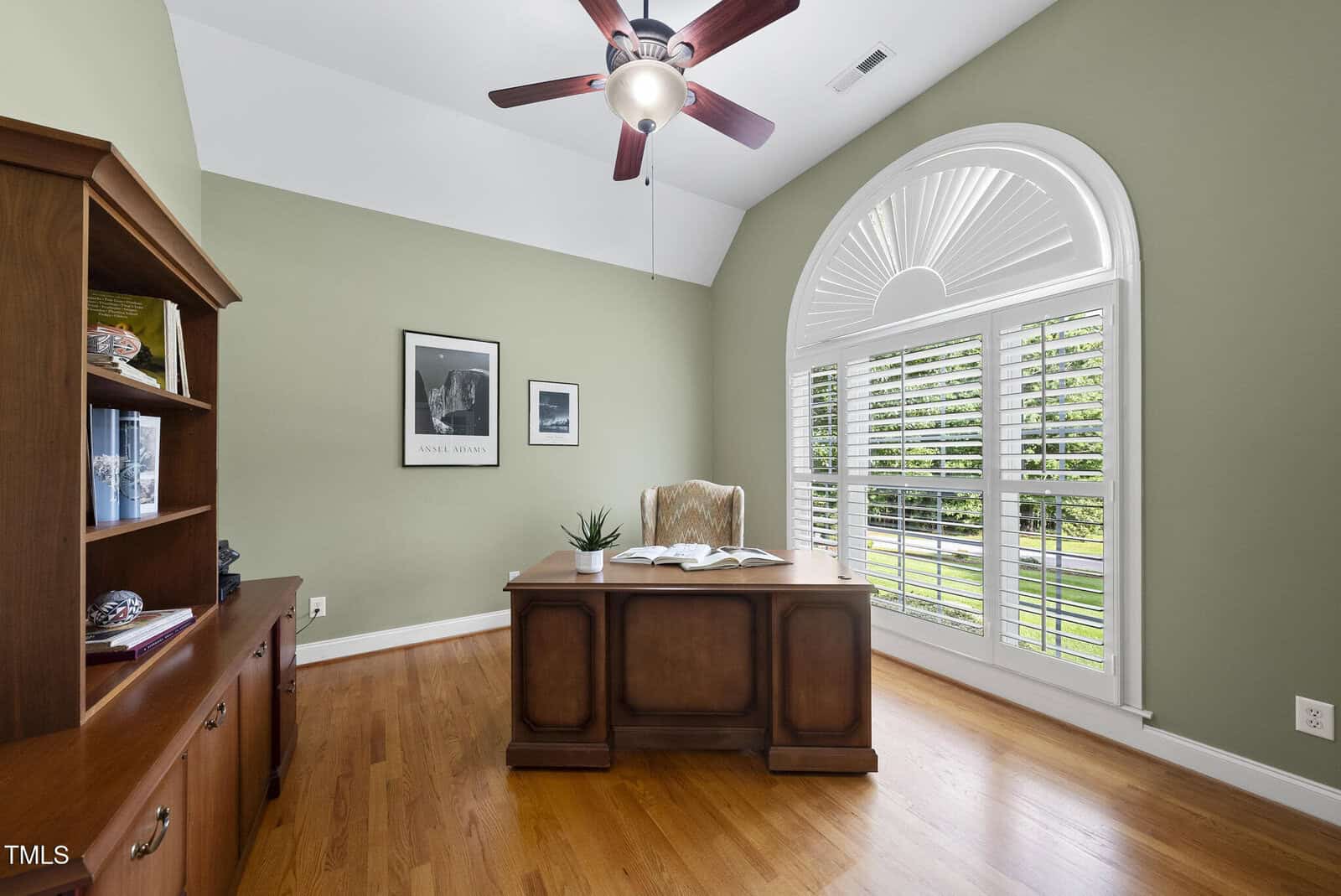 A home office with a wooden desk and chair, built-in shelves, hardwood floors, green walls, a ceiling fan, and a large arched window with white shutters letting in natural light.