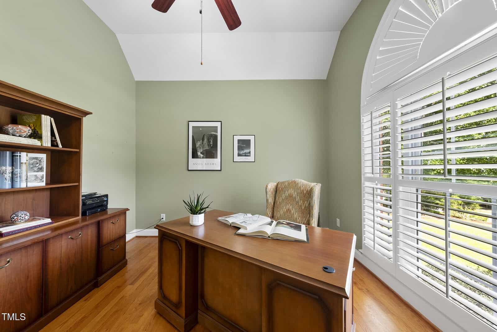 A home office with a wooden desk, an open book, a potted plant, an upholstered chair, a wooden cabinet with shelves, two framed photos on a green wall, and large arched windows with white shutters letting in sunlight.