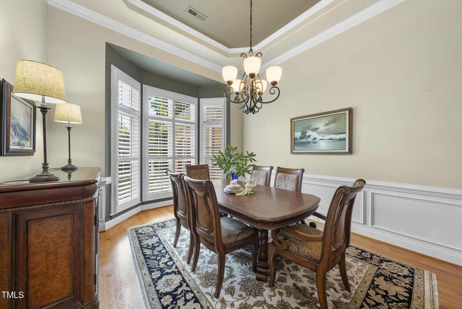 A dining room with a wooden table surrounded by six chairs, a rug underneath, a sideboard with lamps, a chandelier, a plant centerpiece, a painting on the wall, and large windows with white shutters.