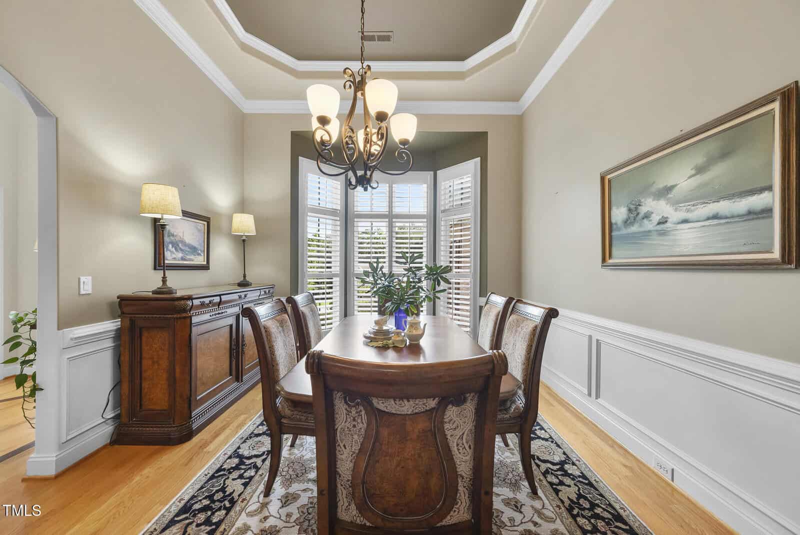 A formal dining room with a wooden table and six chairs, a sideboard with two lamps, a chandelier, a plant centerpiece, an area rug, wainscoting, a large window with shutters, and a framed ocean painting.