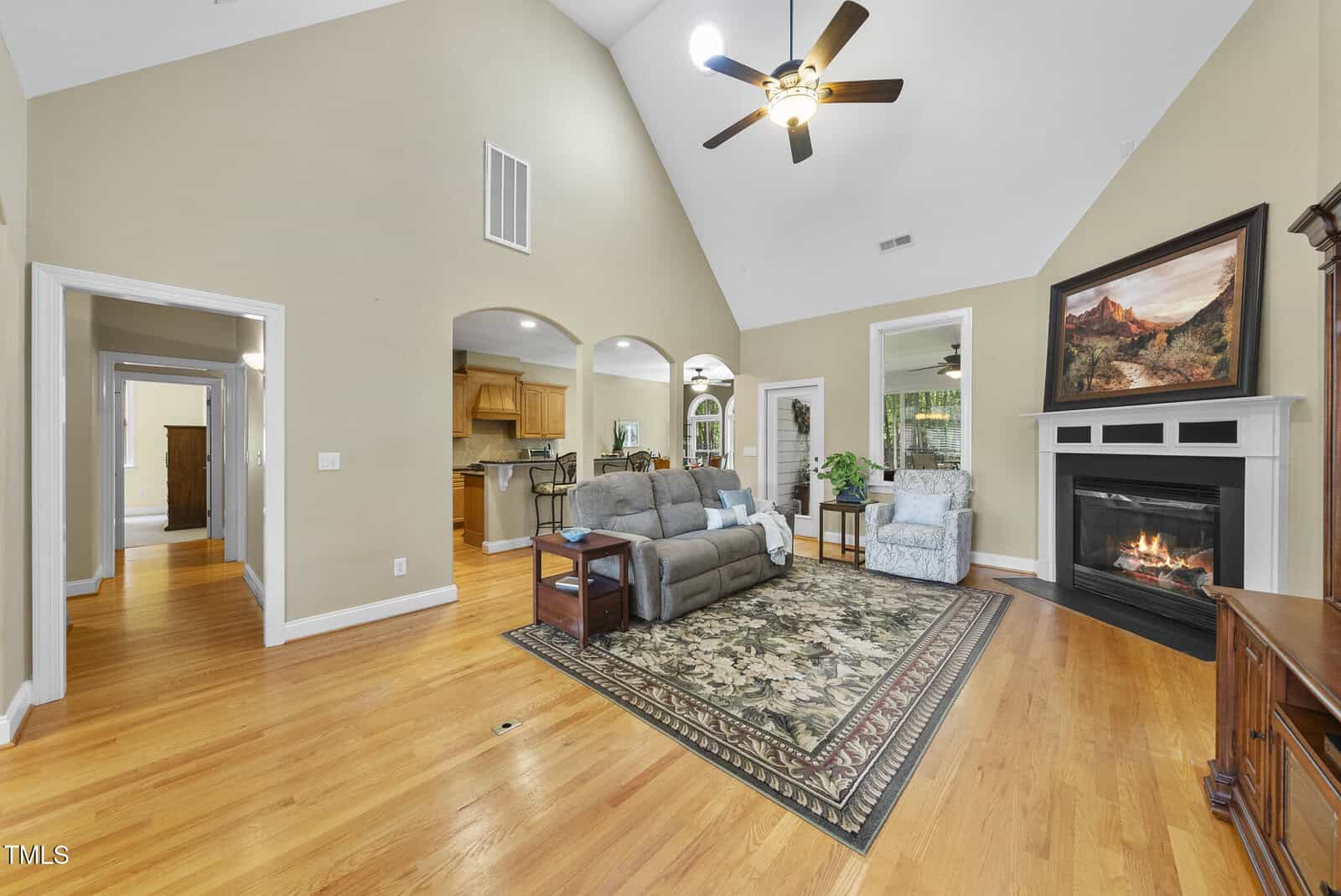 Spacious living room with hardwood floors, vaulted ceiling, gray couch, area rug, ceiling fan, fireplace, armchair, and view into kitchen and dining areas. Light walls and natural light create a warm, inviting atmosphere.