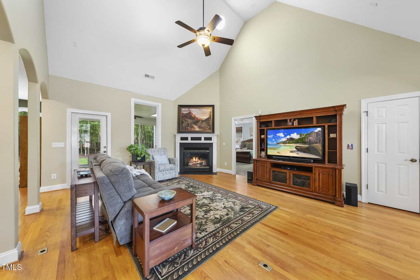 A living room with vaulted ceilings, hardwood floors, a ceiling fan, a fireplace with a painting above it, a large entertainment center with a TV, gray couches, side tables, and a patterned rug.