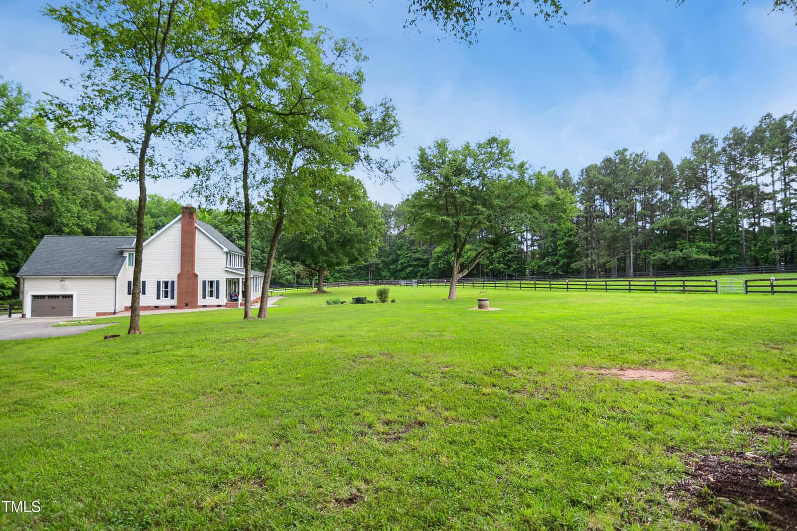 A white house with a brick chimney sits on a large, grassy lawn bordered by trees and a wooden fence. The sky is clear and blue.