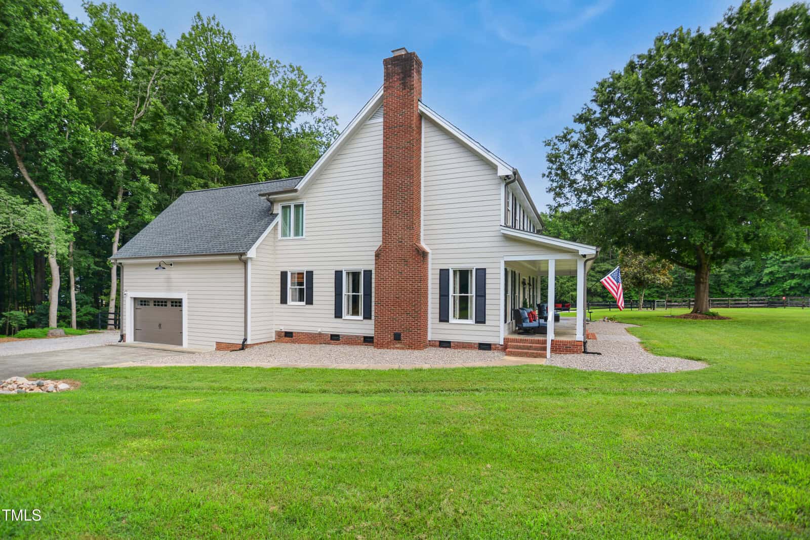Side view of a two-story white house with a prominent brick chimney, a covered porch, black shutters, and an attached garage. The house sits on a well-kept lawn with trees and an American flag in the yard.
