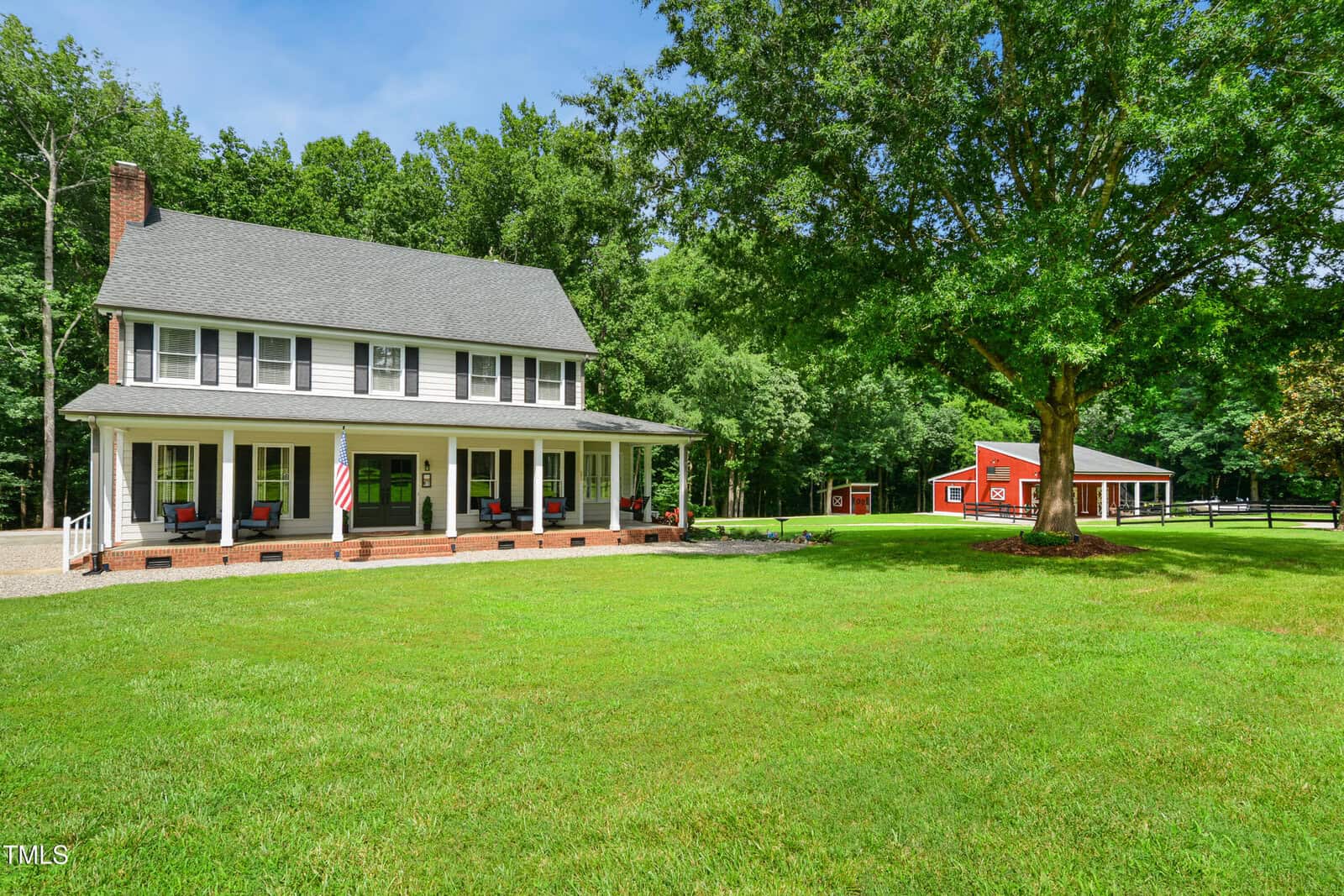 A two-story white house with a covered front porch and black shutters sits on a large, well-kept green lawn. A red barn is visible in the background, surrounded by trees and a wooden fence.