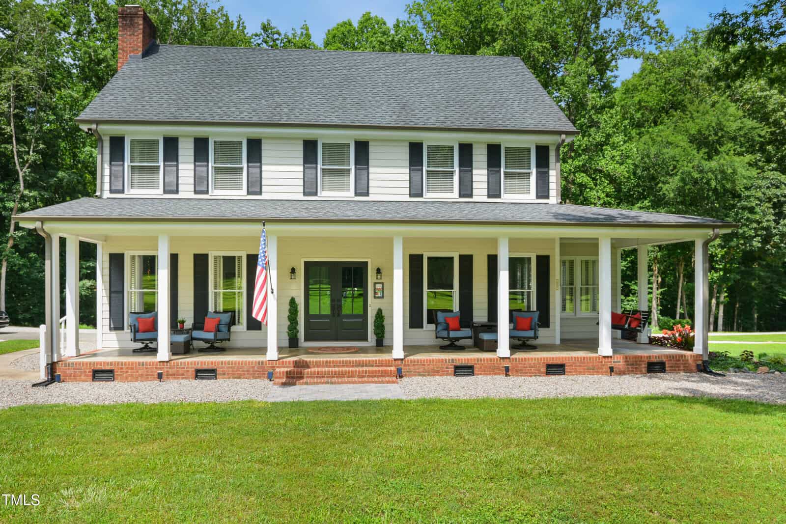 A two-story house with gray siding, black shutters, and a wide wraparound porch featuring seating areas and an American flag. The home is surrounded by lush green grass and tall trees.