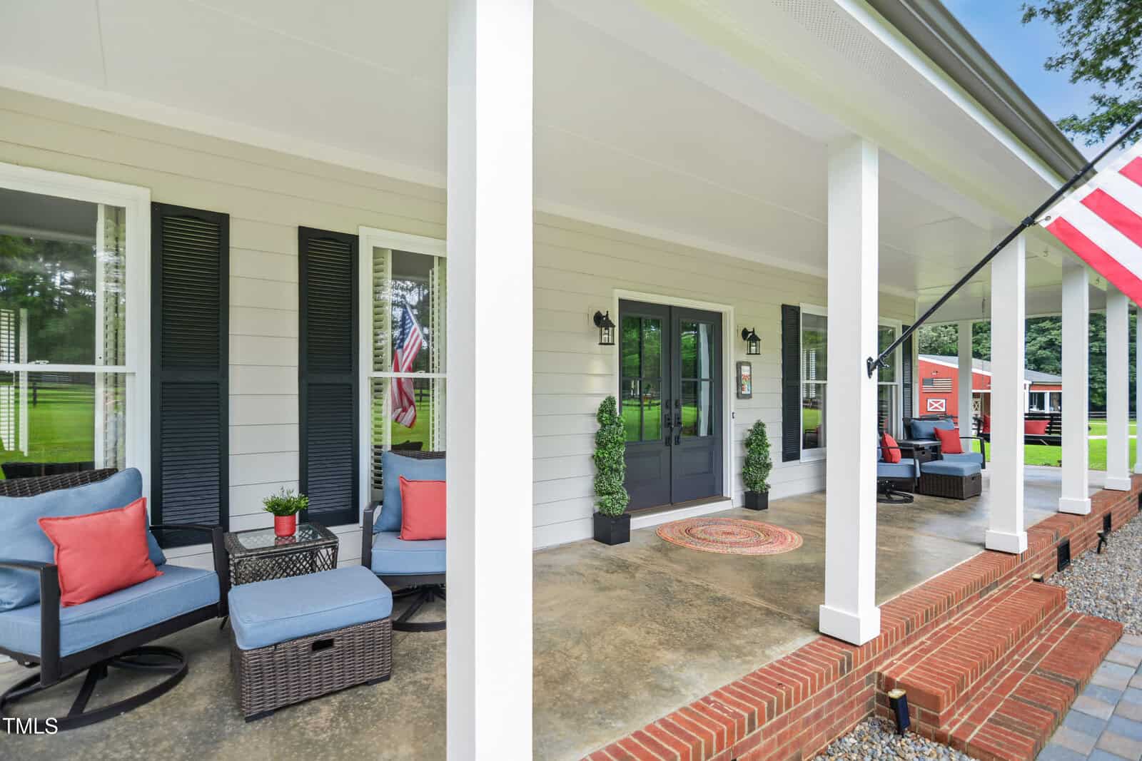 A front porch with white columns, brick steps, dark double doors, potted plants, cushioned chairs with red and blue pillows, and an American flag. A grassy yard and red building are visible in the background.