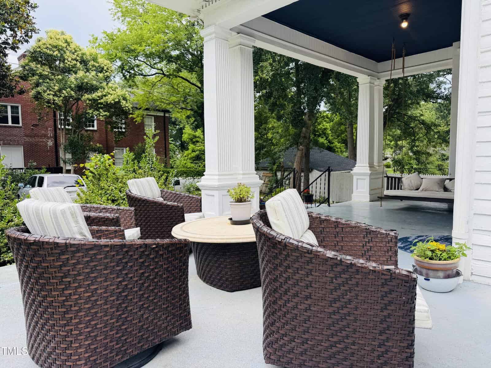 A covered porch with four brown wicker chairs around a round table, potted plants, and a swing hanging in the background, surrounded by white columns and greenery.