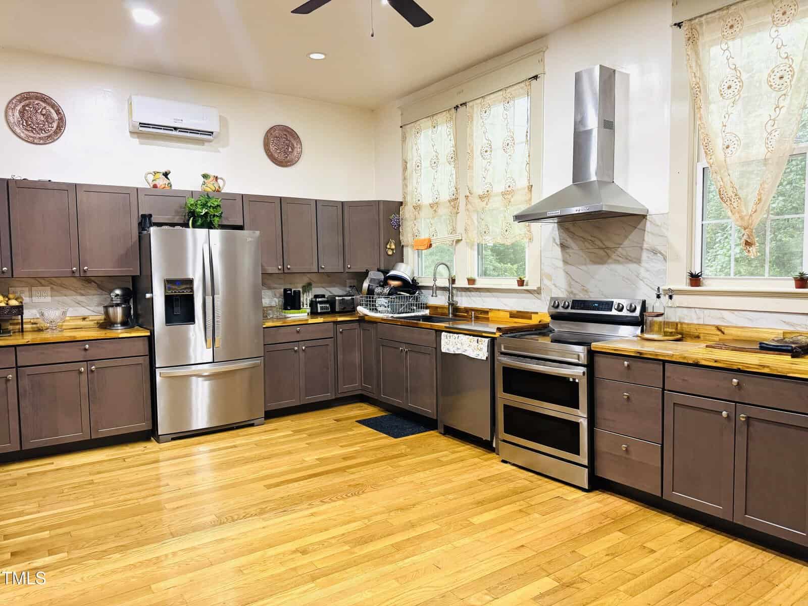 A spacious kitchen with wooden floors, brown cabinets, stainless steel appliances, a double oven, a fridge, a range hood, and countertops with kitchenware. Two large windows have lace curtains.