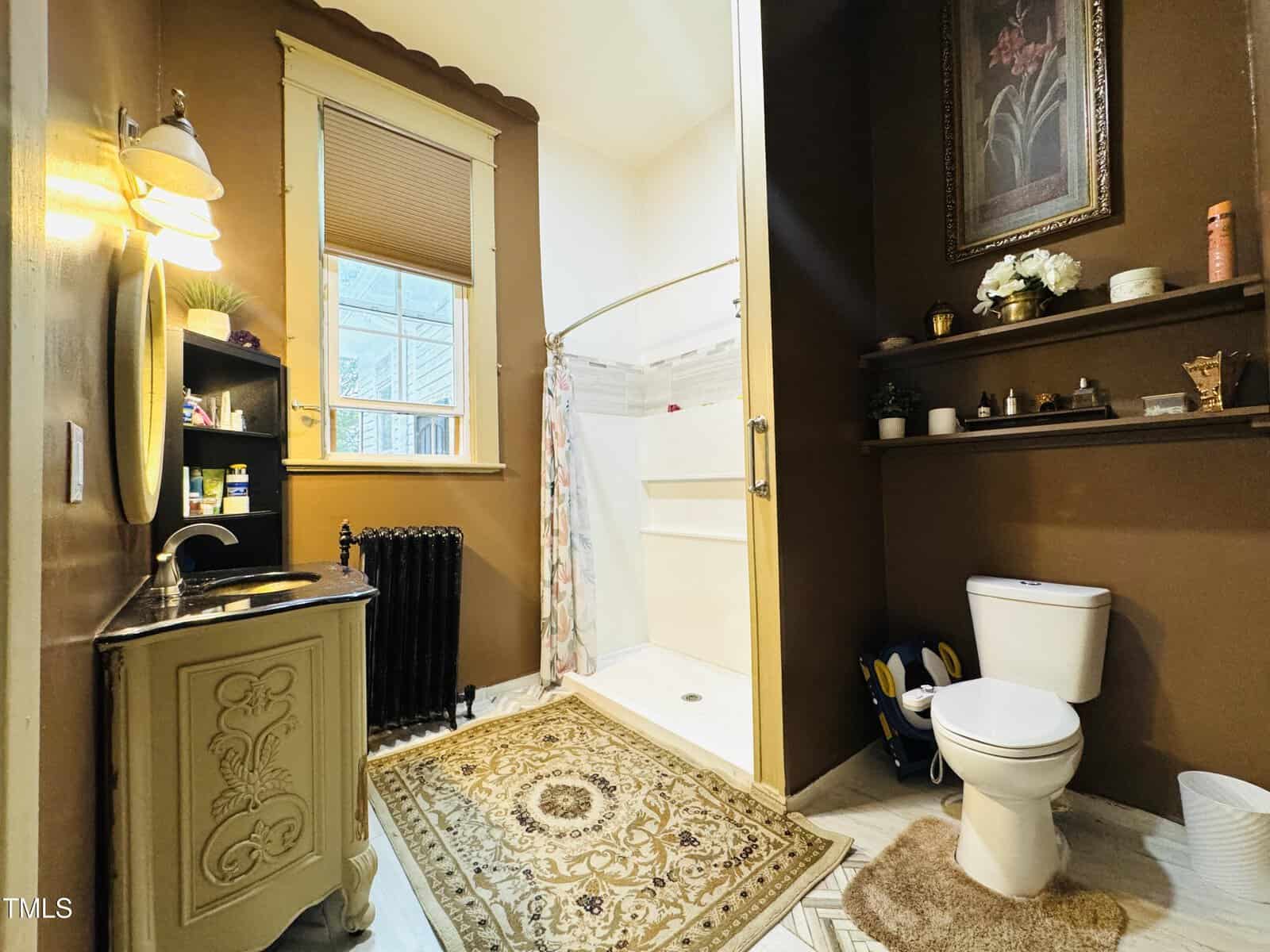 A cozy bathroom with brown walls, a white toilet, a vintage sink, a shower with a curtain, shelves holding plants and toiletries, a window, and a decorative rug on the floor.