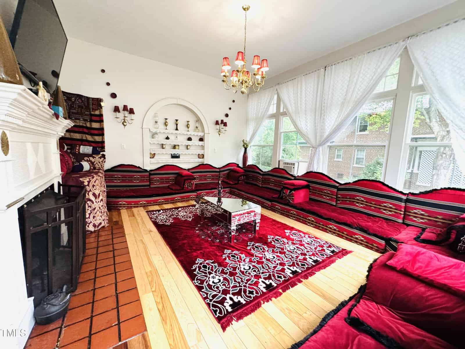 A bright living room with large windows, red and black patterned sectional seating, a red rug, a chandelier, white curtains, and decorative shelves on a white wall.