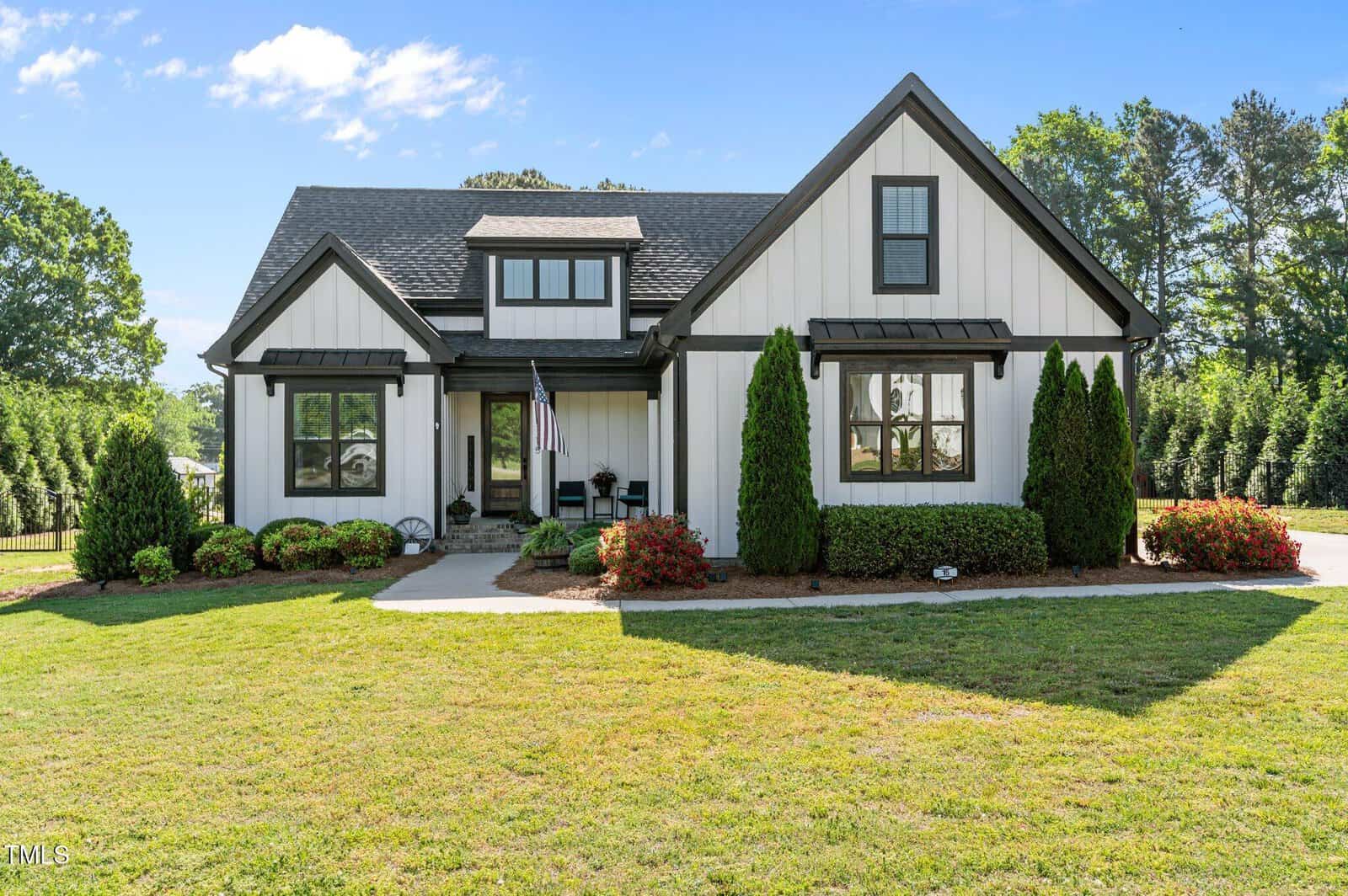 A modern farmhouse with white siding, black trim, and a gabled roof, surrounded by green grass, shrubs, and trees under a blue sky with a few clouds. An American flag hangs by the front porch.