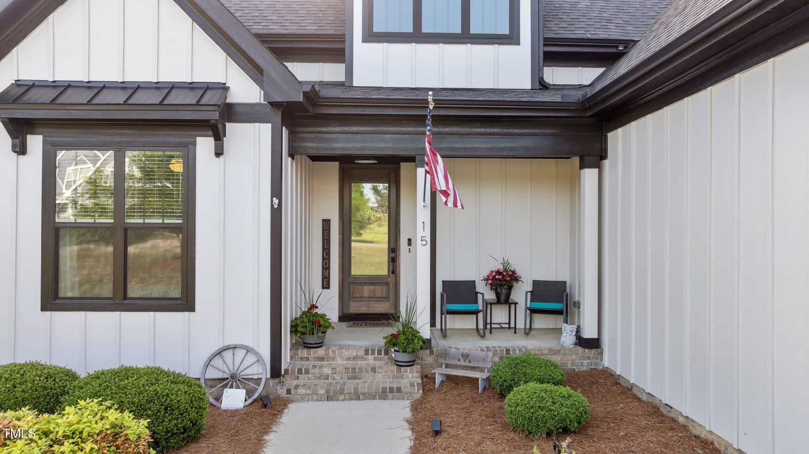 A front porch of a modern house with white panel siding and black trim, an American flag, two black chairs with teal cushions, potted plants, and a glass door with the house number 15.