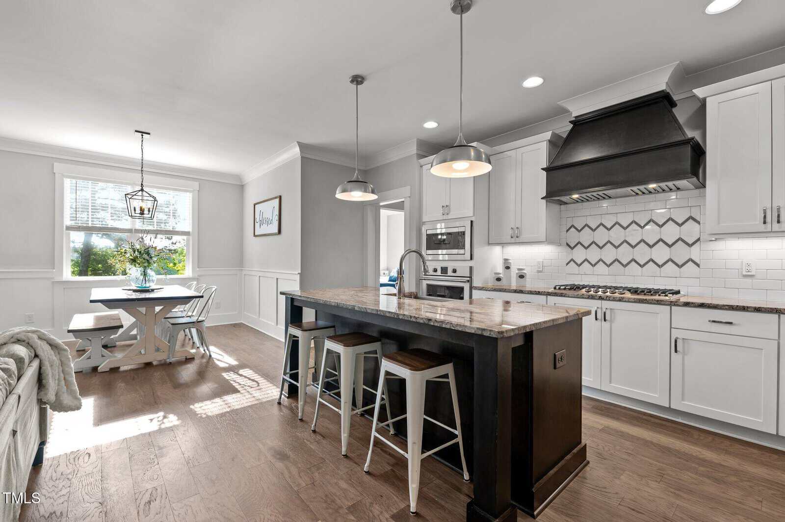 Modern kitchen with a central island featuring three stools, pendant lighting, white cabinetry, stainless steel appliances, and a geometric tile backsplash. A bright dining area with a window and a wooden table is visible in the background.