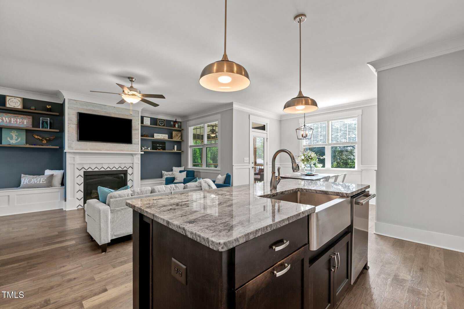 Modern open-concept kitchen and living room with a marble island, farmhouse sink, pendant lights, and wood floors. Living area includes a fireplace, built-in shelves, a wall-mounted TV, and a large window with natural light.