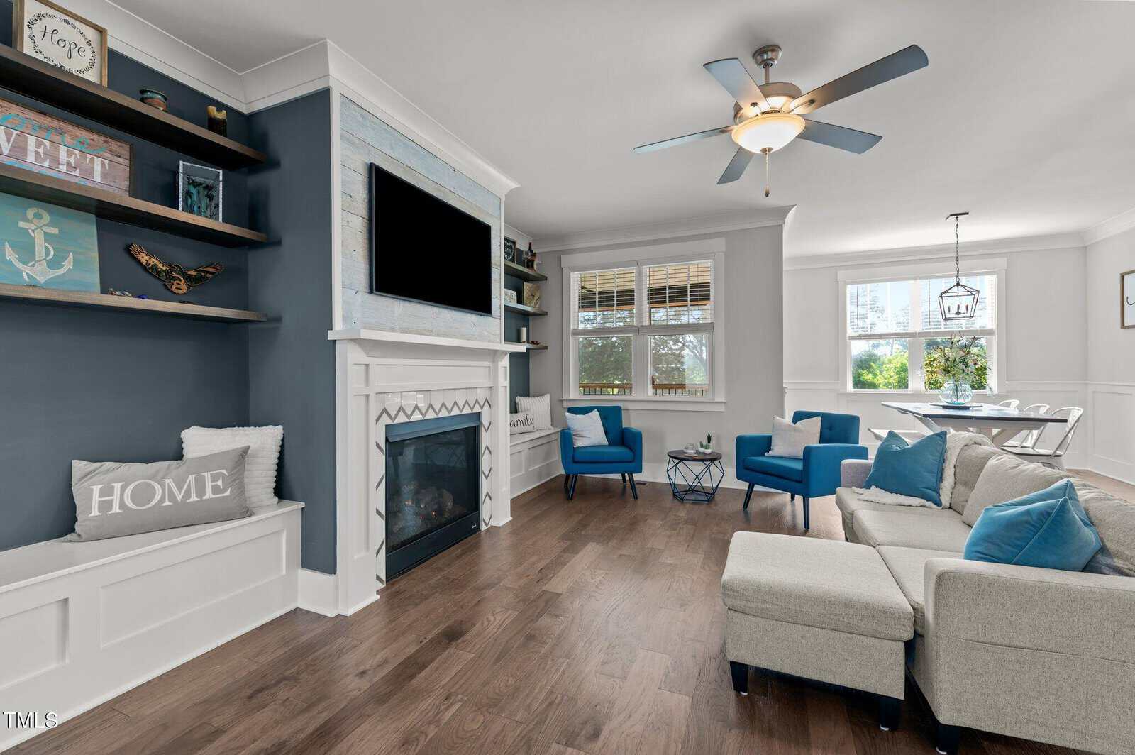 Coastal-themed living room with a fireplace, wall-mounted TV, gray sectional sofa, blue accent chairs, wooden floor, and nautical decor. Large windows let in natural light near a dining table with white chairs.