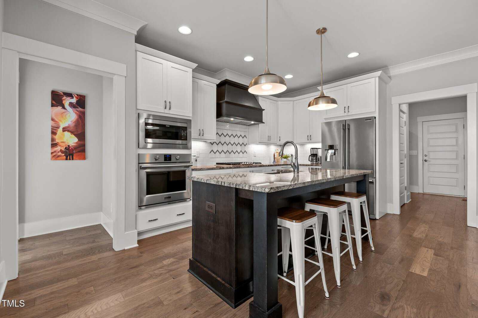 Modern kitchen with white cabinets, stainless steel appliances, a dark island with granite countertop, three stools, pendant lights, wood flooring, and a colorful framed artwork on the left wall.