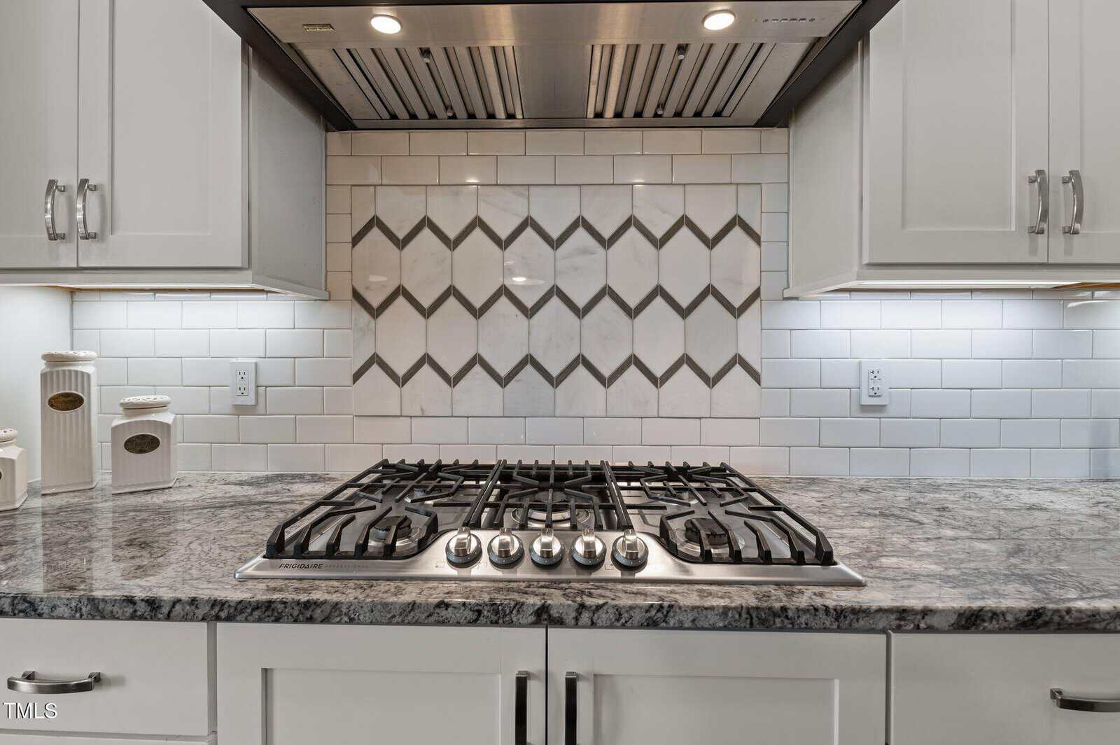 A modern kitchen stove with five gas burners sits beneath a stainless steel range hood. The backsplash features white subway tiles with a geometric accent pattern. White cabinets and marble countertops are also visible.