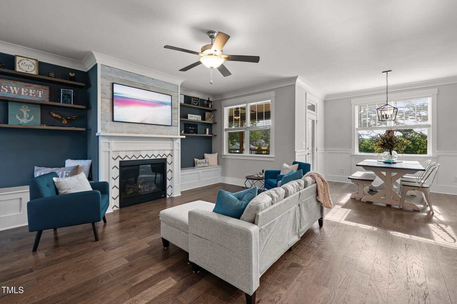A living room with a gray sofa, blue chairs, and a fireplace beneath a wall-mounted TV. Built-in shelves and windows are on either side. Dining area with a white table and chandelier is visible in the background.