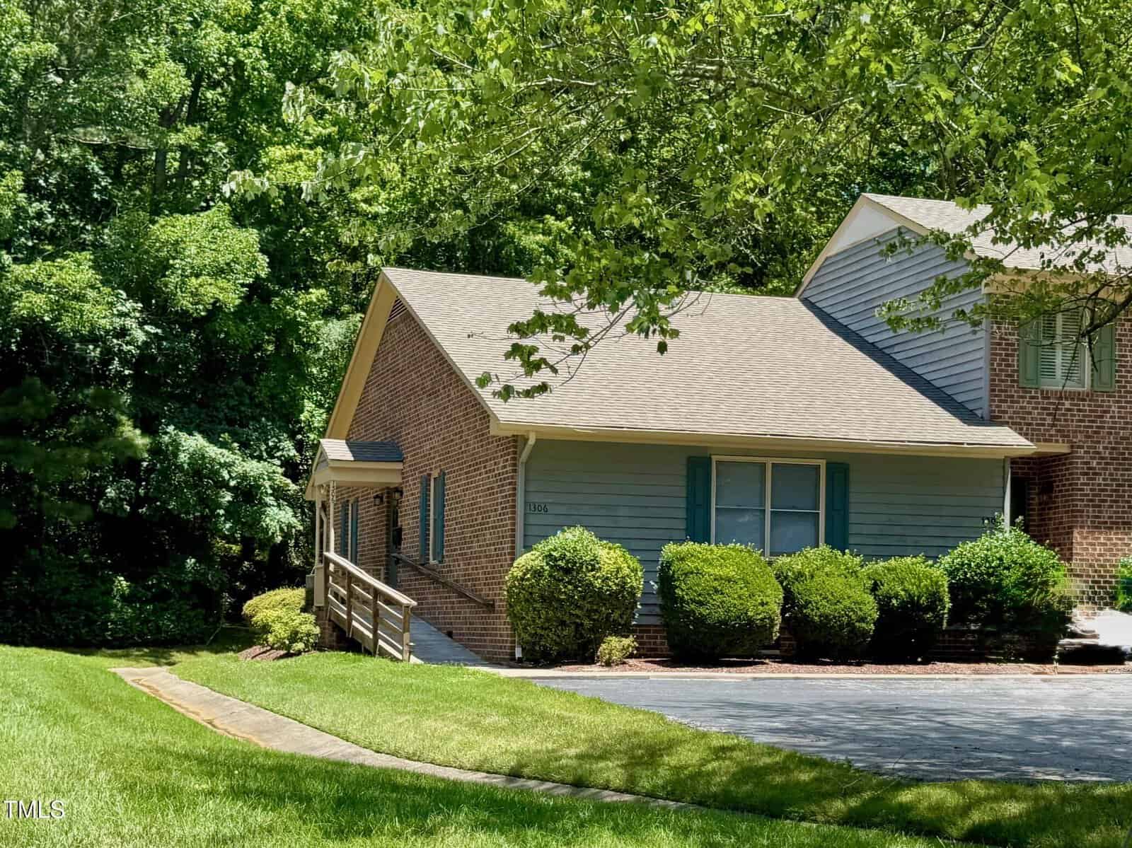 A single-story house at 1306 Oakridge Avenue, Henderson, features blue siding with brick accents, surrounded by neatly trimmed shrubs and trees. A concrete driveway and wooden ramp lead to the front entrance of the well-maintained grassy yard.