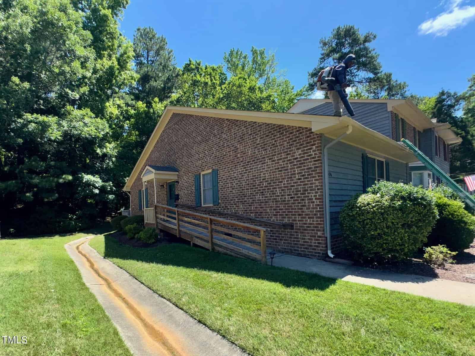 A person wearing safety gear stands near the edge of the roof of a brick house with blue siding, surrounded by green trees, under a clear blue sky. A sidewalk runs along the left side of the house.