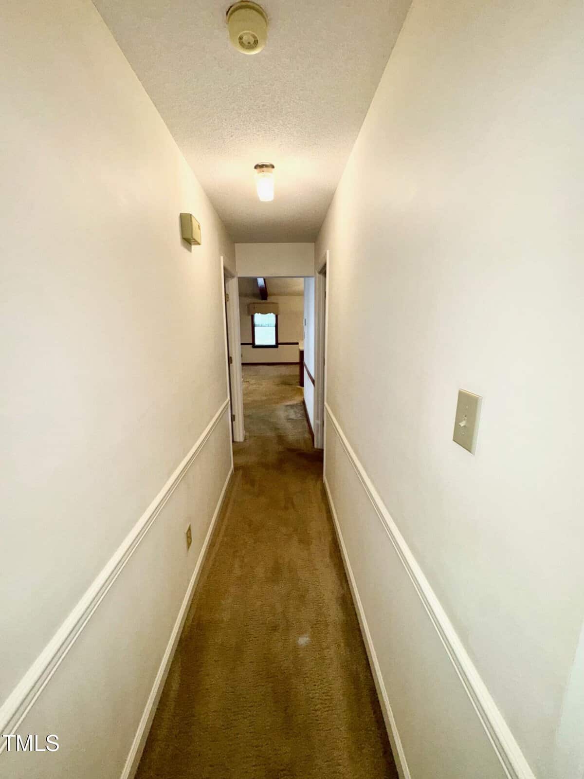 A long, narrow hallway with beige carpet and white walls, lit by two ceiling lights, with doorways on the left and right and a room visible at the end.