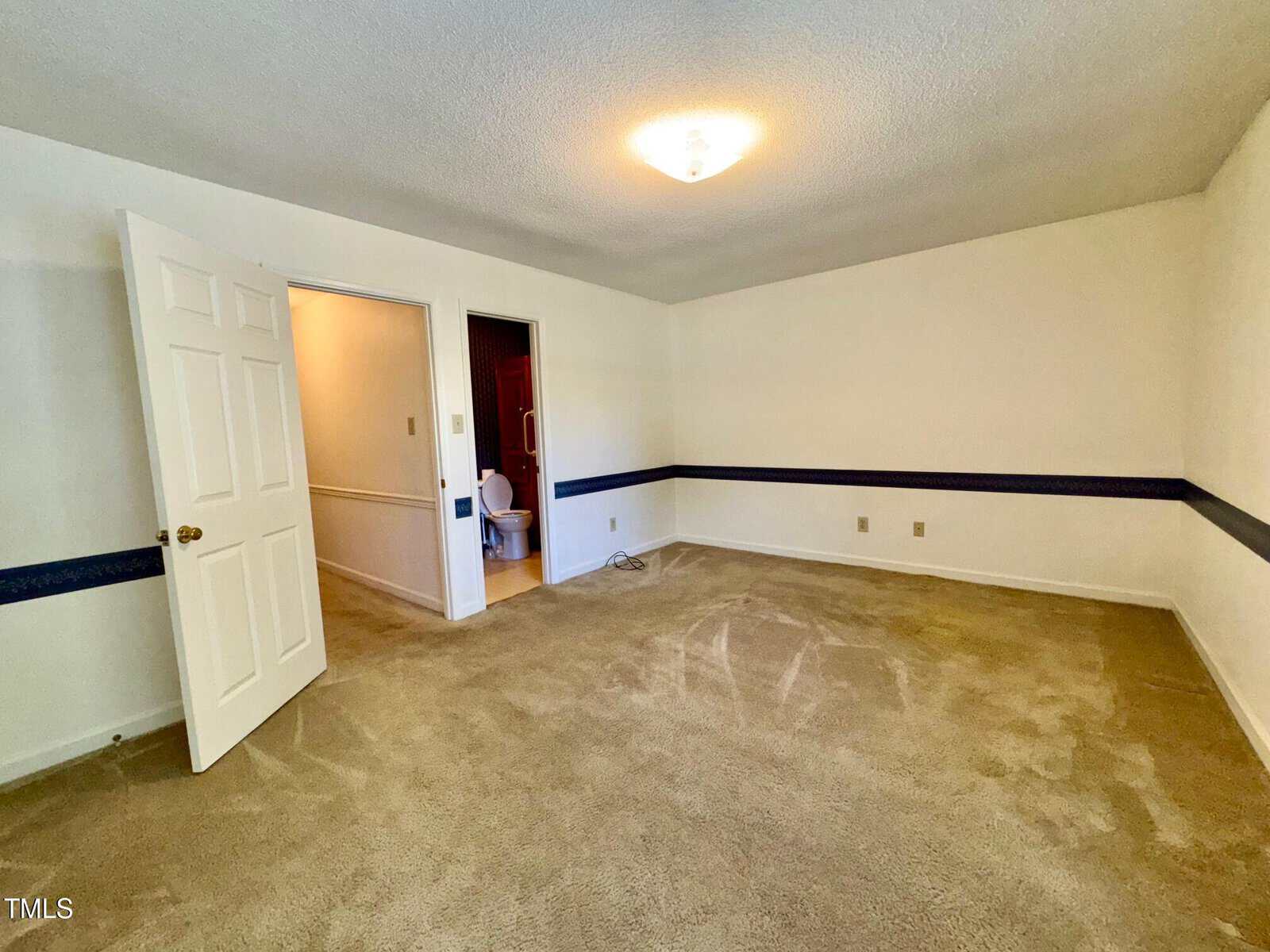 Empty carpeted room with beige walls, a ceiling light, open door leading to a bathroom with a visible toilet, and a dark blue decorative border running around the walls at mid-height.