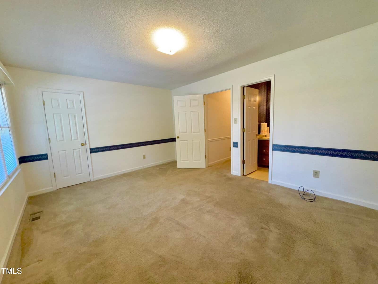 Empty carpeted bedroom with cream walls, two closed doors, an open door to a bathroom, a ceiling light, window on the left, and dark blue wallpaper border around the middle of the walls.