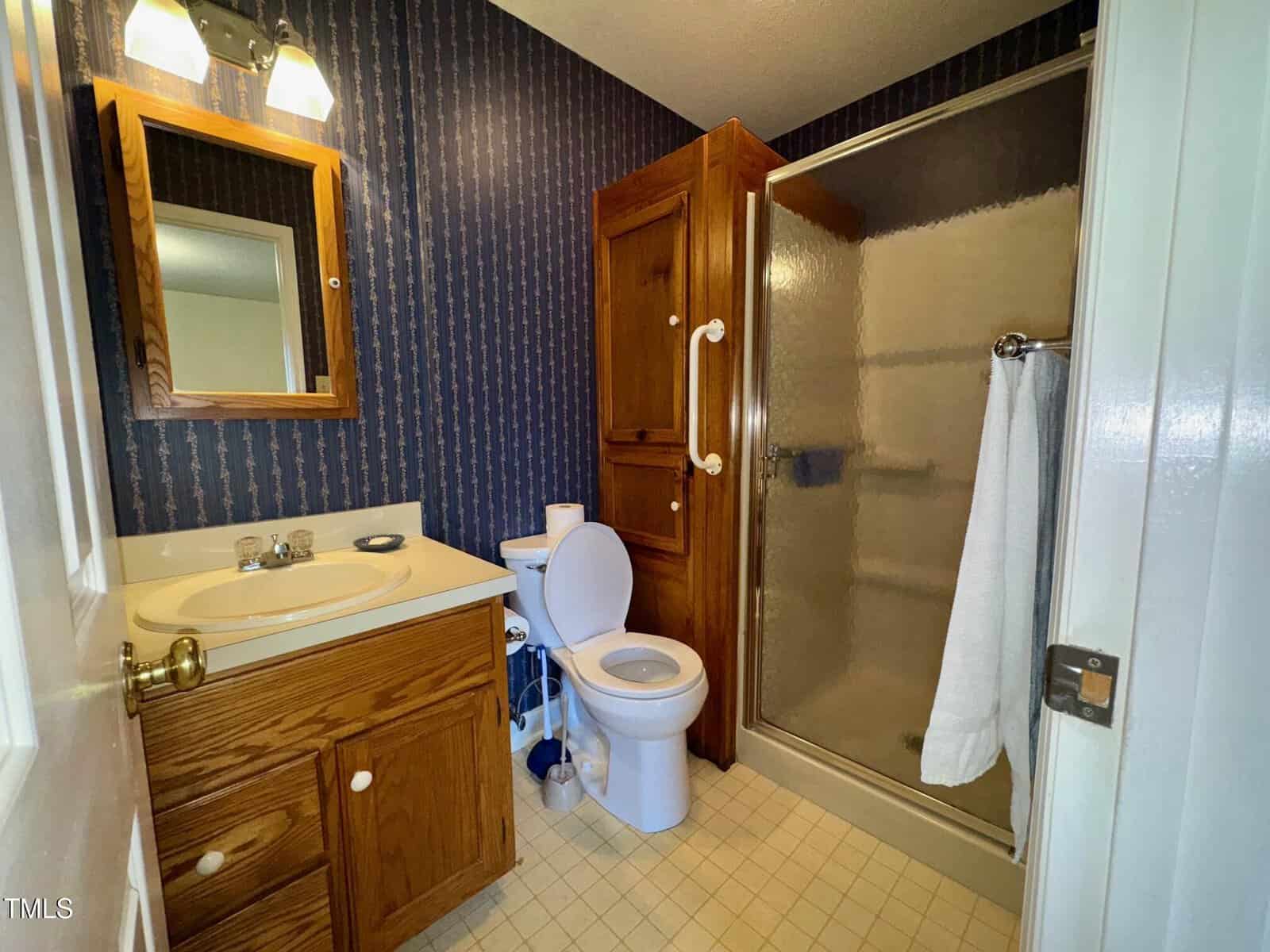 Small bathroom with wood cabinets, a sink, toilet, and a shower with a glass door. A white towel hangs on the shower handle. The wallpaper is dark with vertical patterns, and the floor is covered in light-colored tiles.