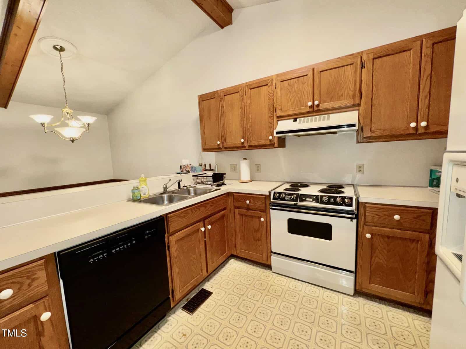 A kitchen with wooden cabinets, a white stove and oven, a white range hood, a dishwasher, a double sink, and beige patterned linoleum flooring. A hanging light fixture is above the sink area.