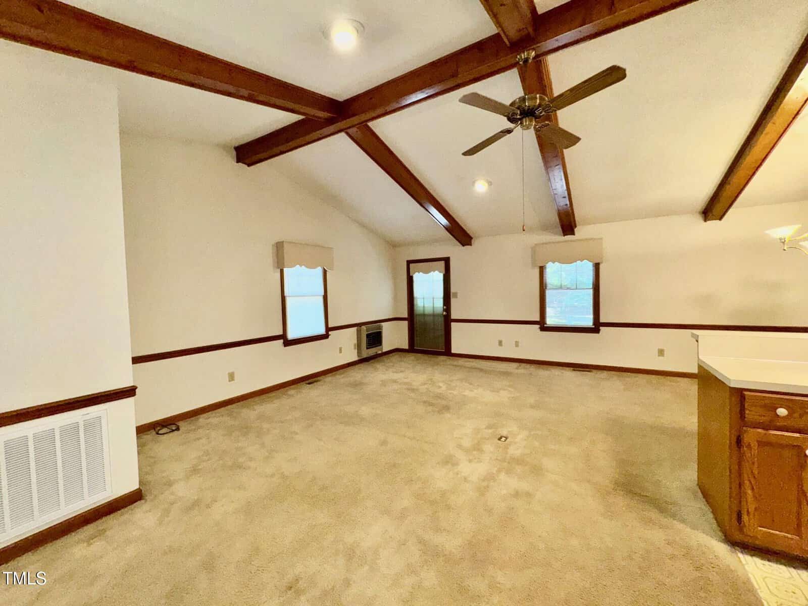 A spacious living room with beige carpet, white walls, exposed wooden ceiling beams, ceiling fan, and several windows. A partial view of a kitchen counter is visible on the right side.