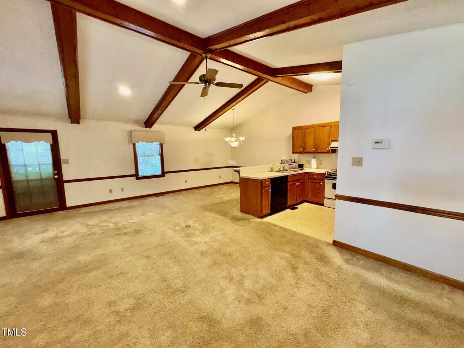 Spacious open-plan living area with vaulted ceiling, exposed wooden beams, carpeted floor, and an adjacent kitchen featuring wood cabinets, a gas stove, and a ceiling fan. Large window and door let in natural light.