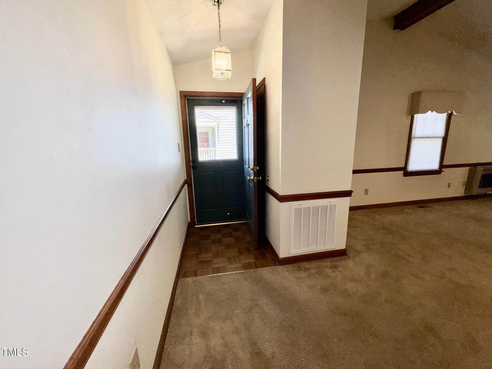 A small entryway with a black front door that is open, leading into a carpeted living room. The walls are white with wooden trim, and a ceiling light hangs above the entry. A window is visible through the door.
