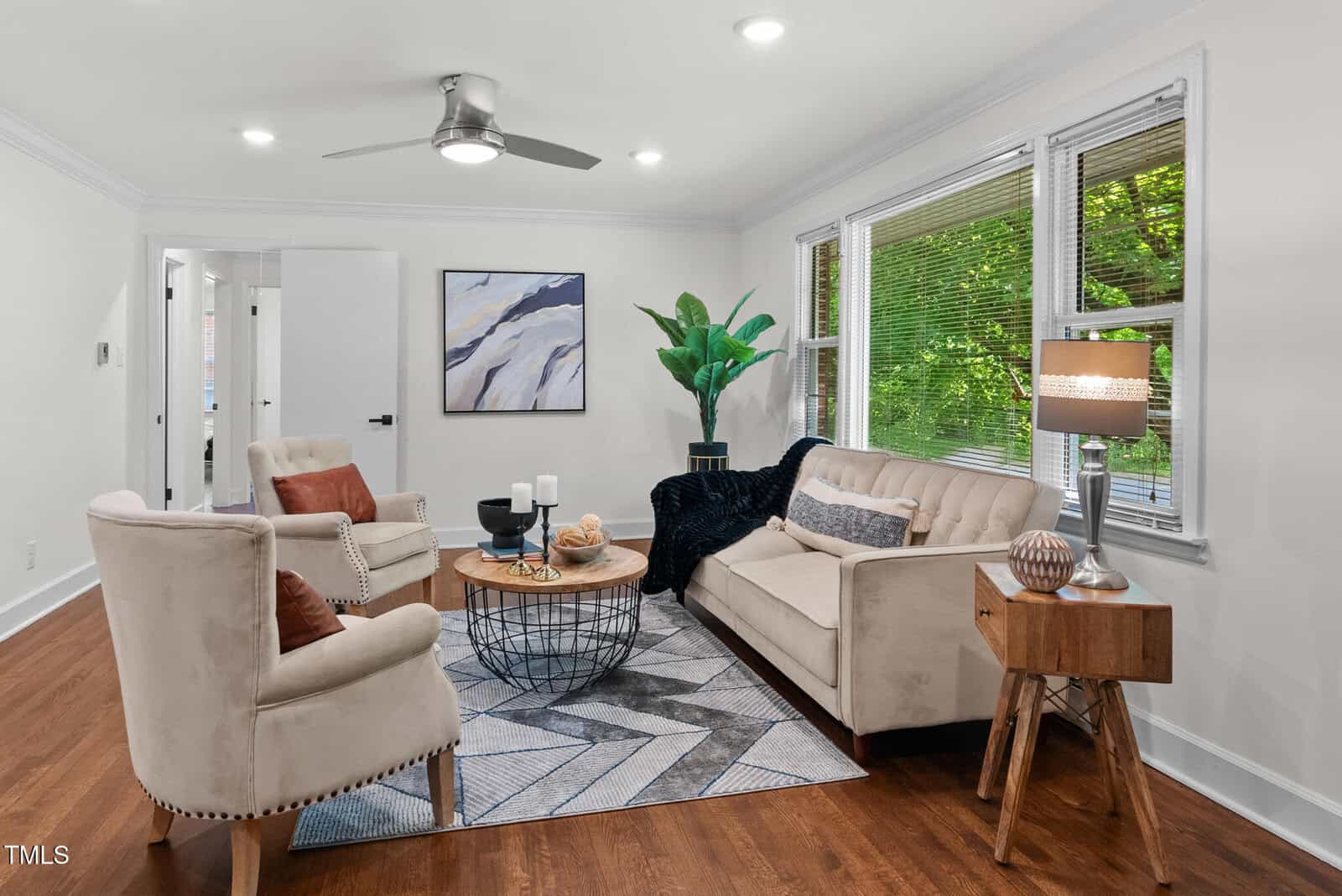 A modern living room with two beige armchairs, a matching sofa, a round wooden coffee table, a geometric rug, a side table with a lamp, a large plant, wall art, hardwood floors, and large windows with blinds.
