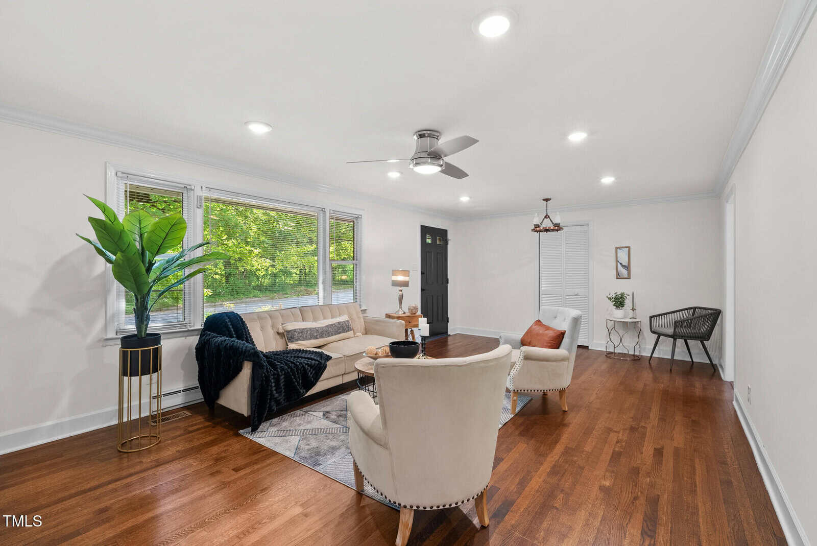 Bright living room with hardwood floors, beige sofas and chairs, a black front door, large window with greenery outside, indoor plant, area rug, and a small desk with a chair in the back corner.