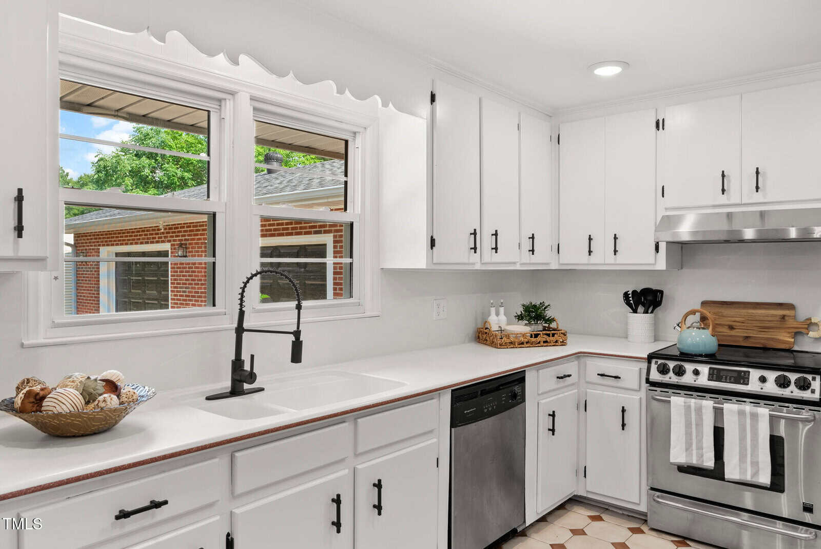 Bright kitchen with white cabinets, black hardware, stainless steel appliances, a farmhouse sink with a black faucet, large window, and a bowl of decorative items on the counter. Neutral decor and hexagon tile flooring.