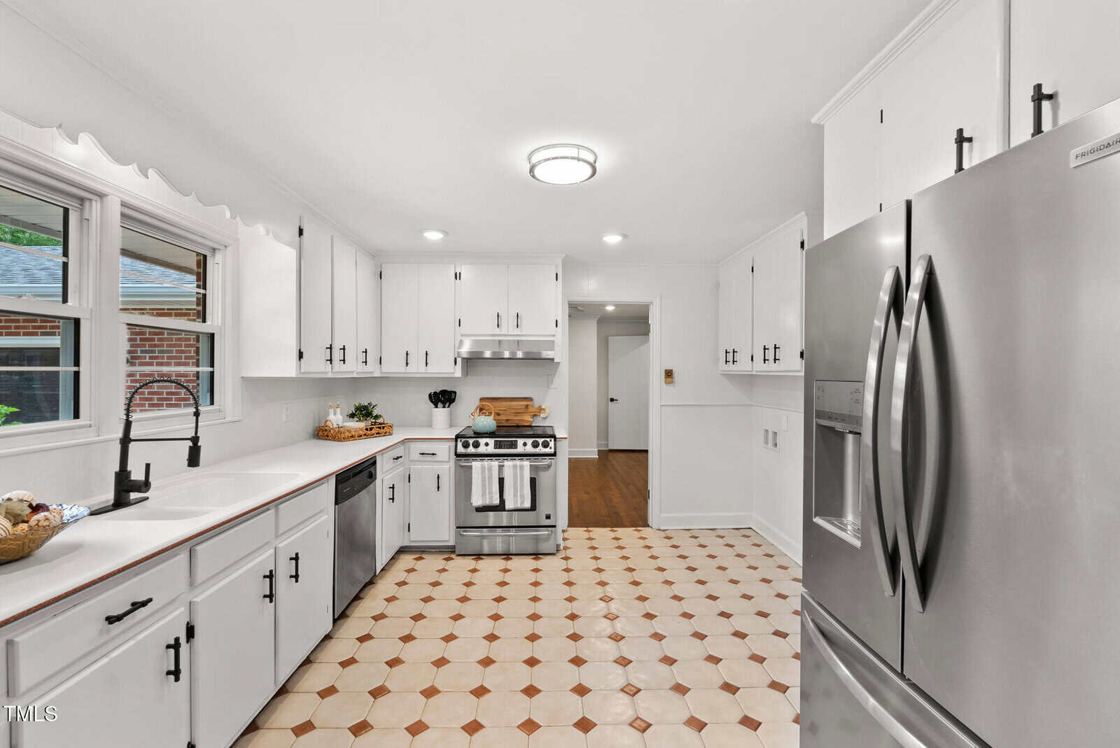 Bright kitchen with white cabinets, stainless steel appliances, double-door refrigerator, a gas stove with oven, black faucet, large window over the sink, and a patterned tile floor in a neutral color scheme.