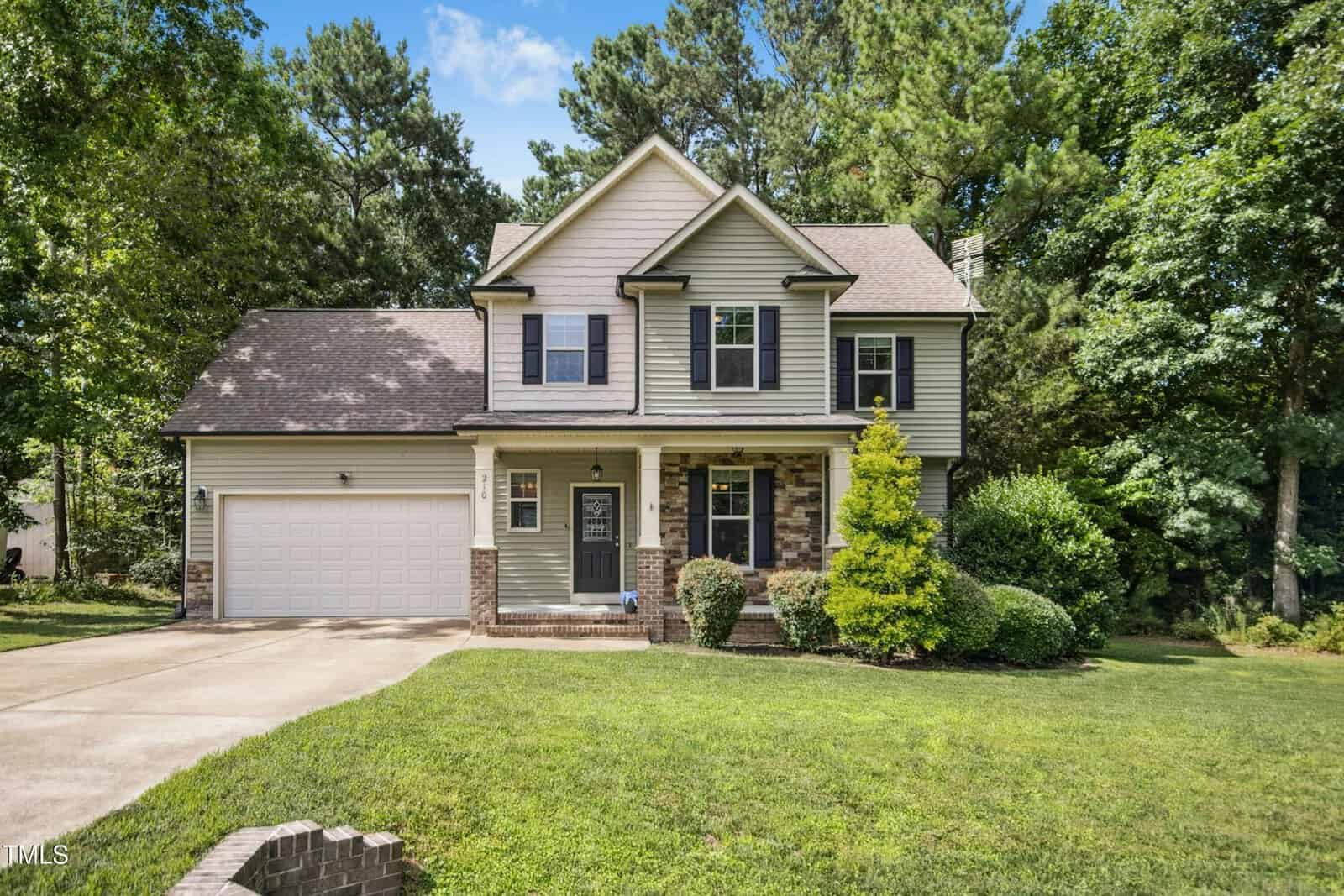 Two-story suburban house with gray siding, brick accents, attached two-car garage, and a covered front porch. The house is surrounded by a well-kept lawn, shrubs, and tall trees under a blue sky.