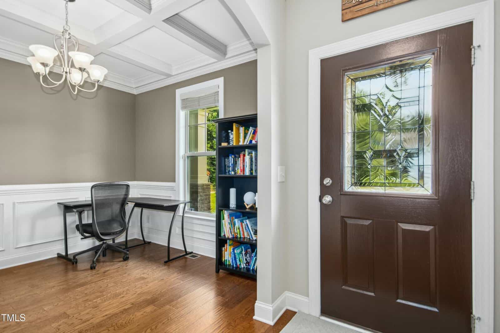Home office with a black desk and chair by a window, a bookshelf filled with books, hardwood floors, beige walls with white wainscoting, and a brown front door with a decorative glass window.