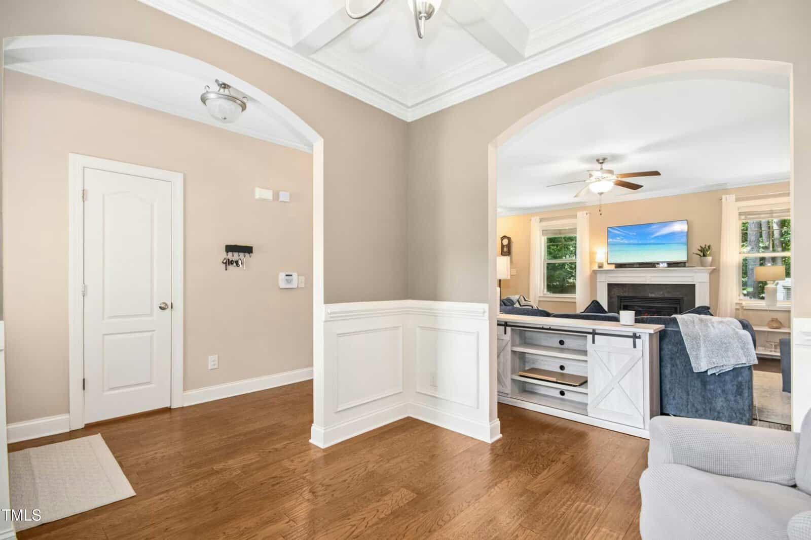 A bright entryway with hardwood floors leads to a living room through an arched doorway. The living room has a fireplace, mounted TV, windows, and ceiling fan. Neutral walls and white trim give the space an airy feel.