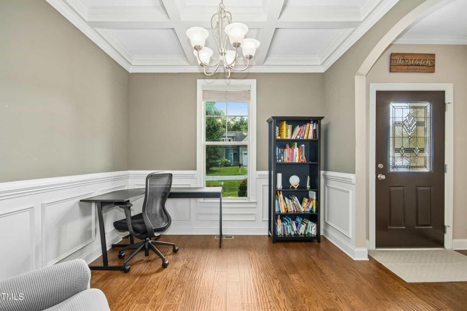 A home office with a black desk and chair, a black bookshelf filled with books, a window showing greenery outside, wooden floors, wainscoting, and a dark front door with decorative glass.